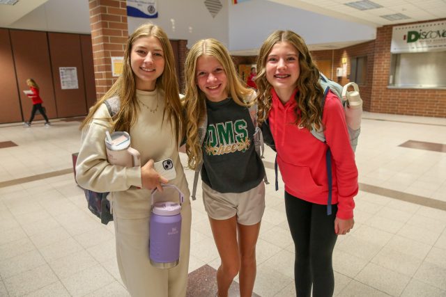 three middle school aged female students standing in school hallway with backpacks and insulated cups