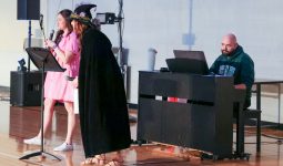 Two people performing on stage in front of a piano, one person is holding a microphone and the other is wearing a witch