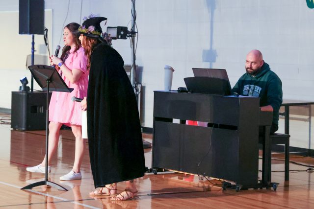 Two people performing on stage in front of a piano, one person is holding a microphone and the other is wearing a witch