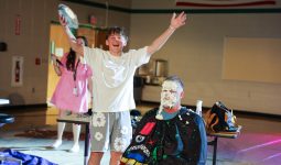 In a school gymnasium, a group of people are celebrating an event, with one person holding a trophy and another wearing a