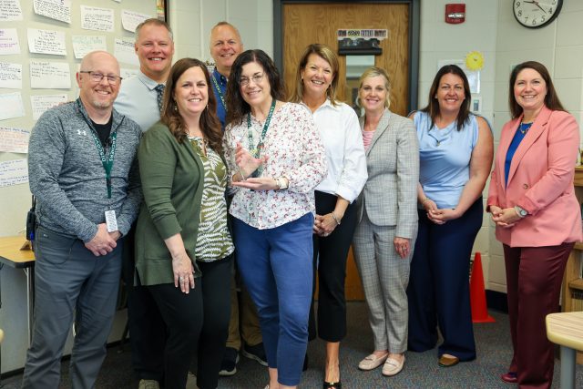 A group of people posing together in a classroom setting, likely educators or staff members, standing in front of a bulletin