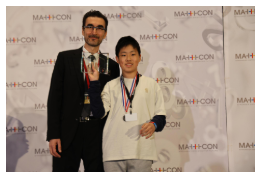 Two people standing on stage at an event. A man in a suit holding a plaque and a boy in a white shirt wearing a medal around