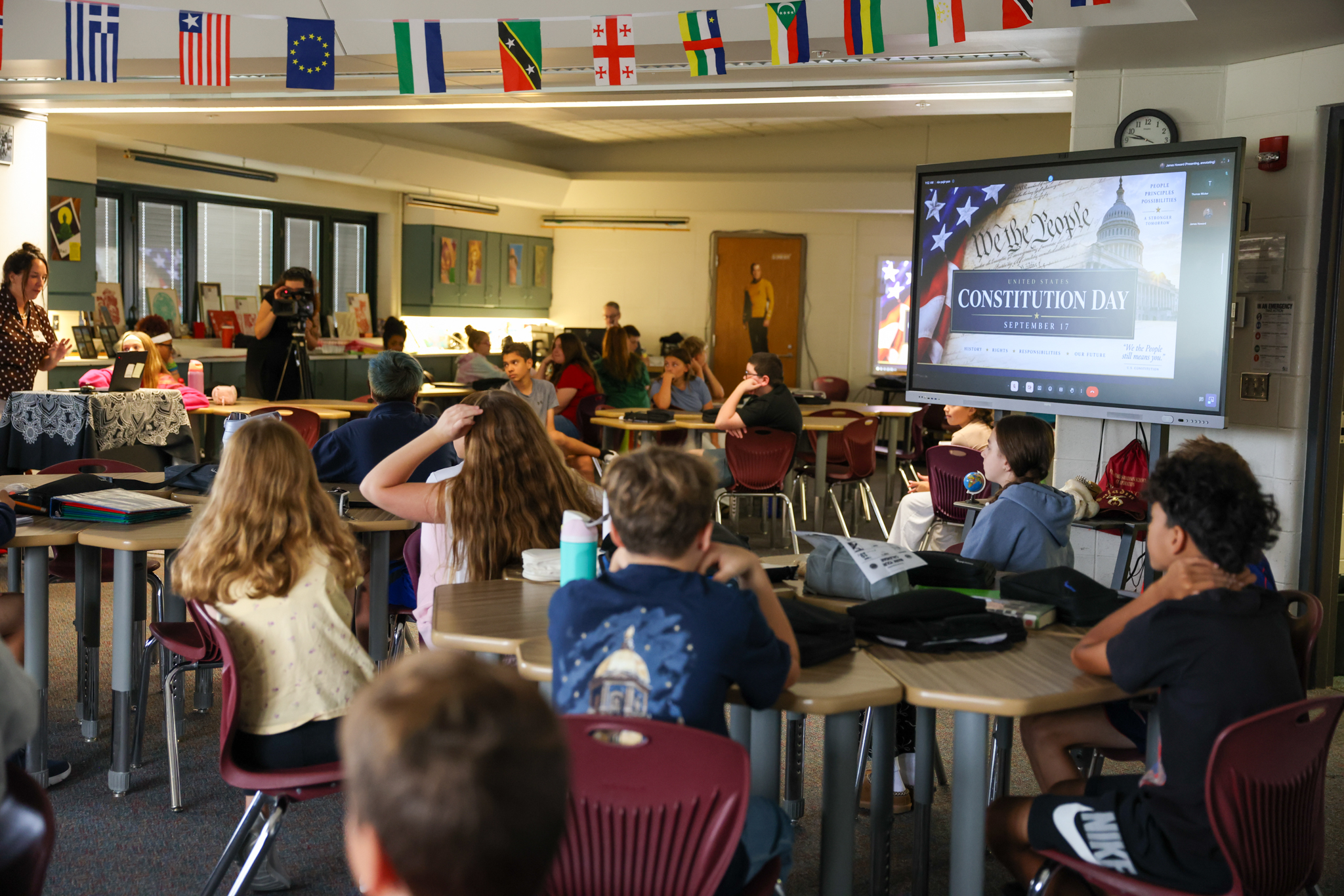 A classroom setting with students seated at desks and a teacher presenting at the front.