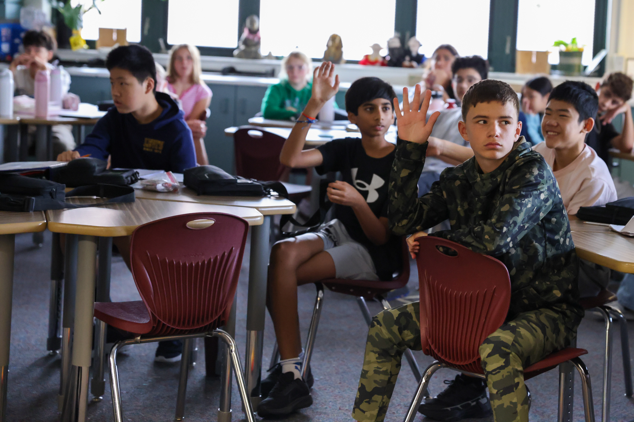 A classroom scene with a young man in camouflage clothing waving from the front row, sitting at a desk with other students,