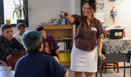 A woman in a classroom, gesturing towards the camera, with a group of students attentively watching her.