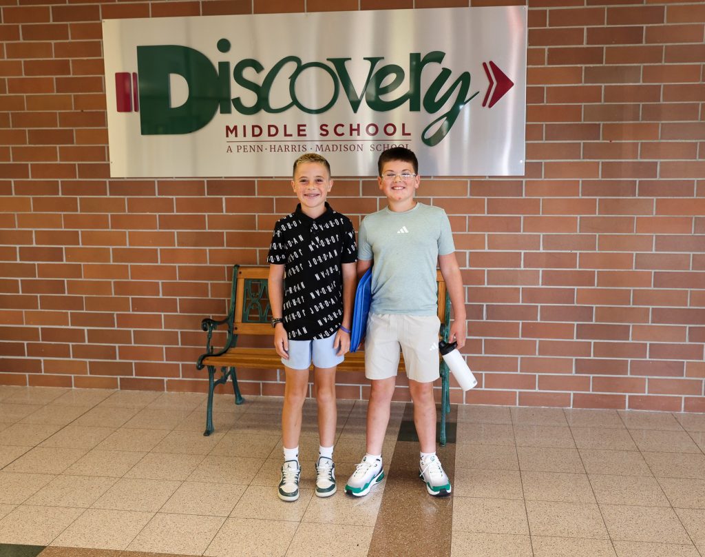 Two middle school aged boys standing in front of a Discovery Middle School sign in the school's hallway.