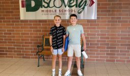 Two middle school aged boys standing in front of a Discovery Middle School sign in the school's hallway.